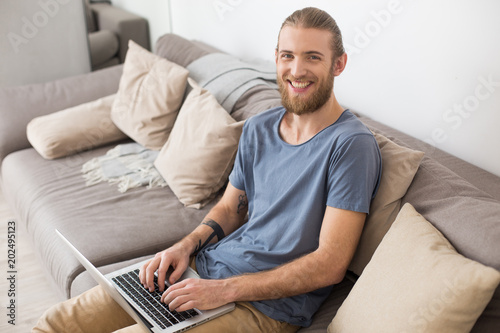 Portrait of young smiling man sitting on big gray sofa with laptop and happily looking in camera isolated