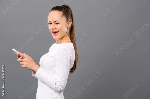 Young and beautiful. Cheerful positive woman winking to you while holding her smartphone