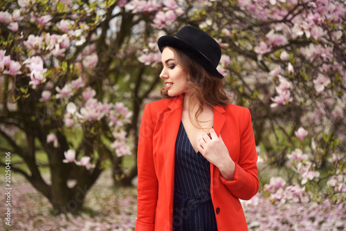 Spring fashion girl outdoors portrait in blooming trees. Beauty Romantic woman in flowers. Sensual Lady enjoying Nature. Model in casual hat , trendy red jacket in orchard smile. Long cerly hair.