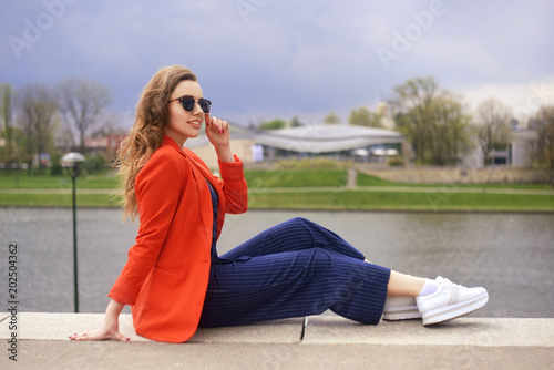Beautiful girl wearing sunglasses at riverside. Girl sitting on pier and lookingat the river. Beautiful young girls outdoors in Sunny weather. Fashion model travel. Girl with long hair in red jacket.