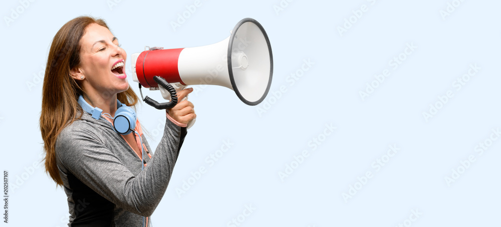 Middle age gym fit woman with workout headphones communicates shouting loud holding a megaphone, expressing success and positive concept, idea for marketing or sales isolated blue background Middle age gym fit woman with workout headphones communicates shouting loud holding a megaphone, expressing success and positive concept, idea for marketing or sales isolated blue background