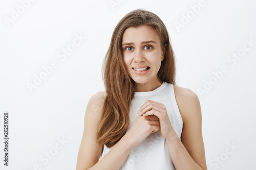 Oops, very awkward situation. Portrait of emberrassed good-looking european woman, rubbing hands over chest and grimacing, feeling ashamed and guilty, feeling anxious and nervous over gray wall photo