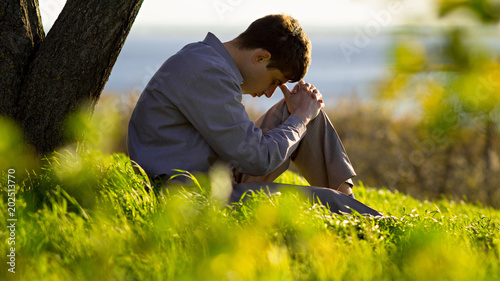 Fotografie, Obraz young man praying to God in the nature bowing his head to his knees, concept rel