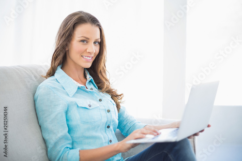 Smiling brunette sitting on sofa using laptop