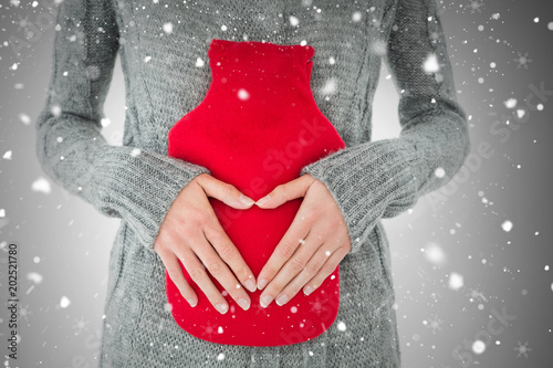 Mid section of a woman holding red hot water bag on abdomen against snow falling photo