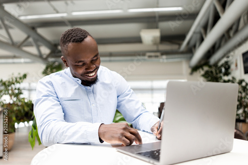 Photos Smiling African man sitting in an office behind a Desk making notes in a noteboo