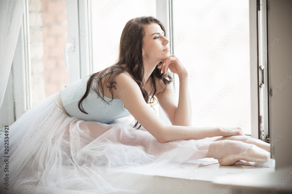 Beautiful ballerina girl is sitting on windowsill in studio