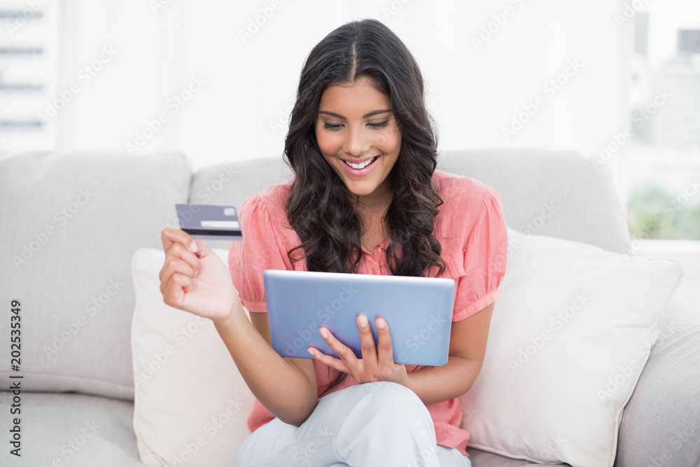 Smiling cute brunette sitting on couch holding credit card and tablet Smiling cute brunette sitting on couch holding credit card and tablet