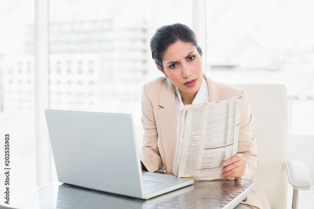 Stern businesswoman holding newspaper while working on laptop looking at camera Stern businesswoman holding newspaper while working on laptop looking at camera