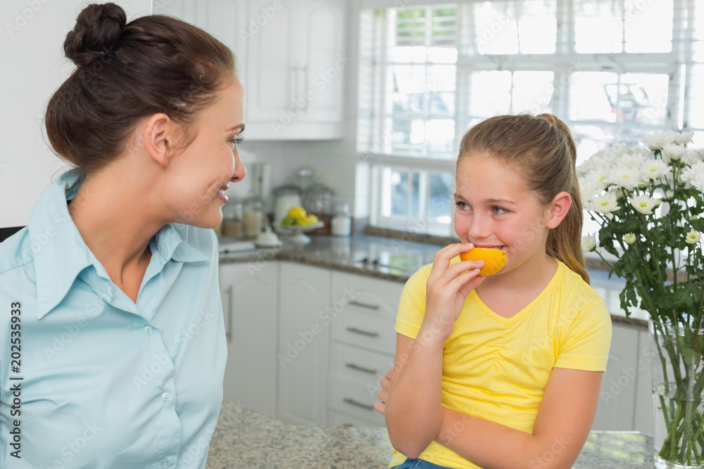 Mother looking at daughter eating orange in kitchen