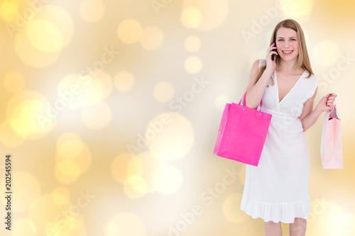 Happy blonde holding shopping bags and talking on phone against yellow abstract light spot design