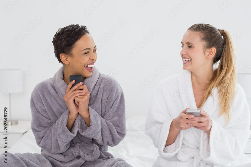 Cheerful friends in bathrobes text messaging on bed Cheerful friends in bathrobes text messaging on bed