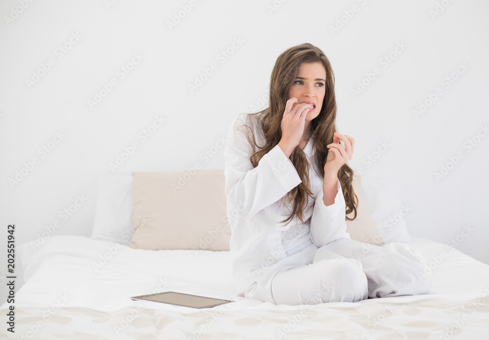 Worried casual brown haired woman in white pajamas sitting on her bed Worried casual brown haired woman in white pajamas sitting on her bed