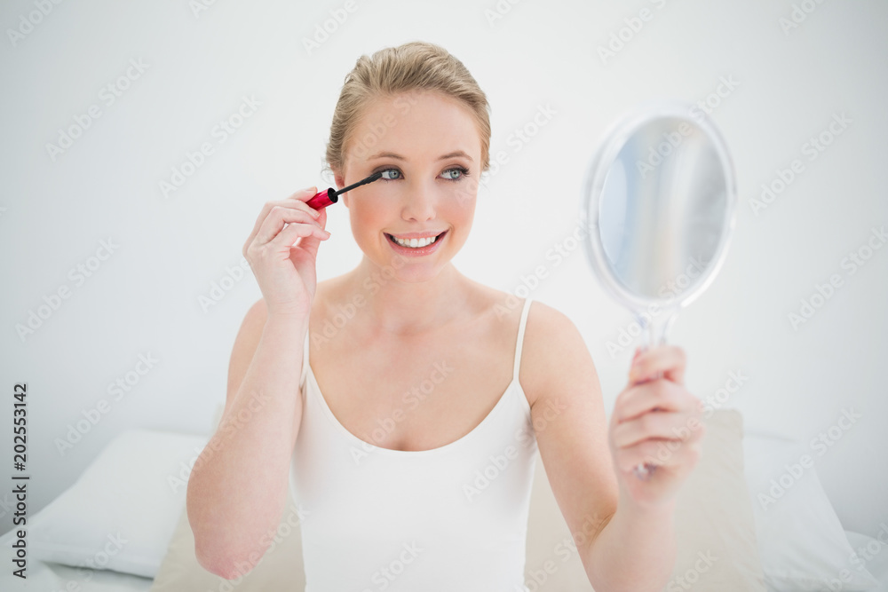 Natural cheerful blonde holding mirror and applying mascara Natural cheerful blonde holding mirror and applying mascara