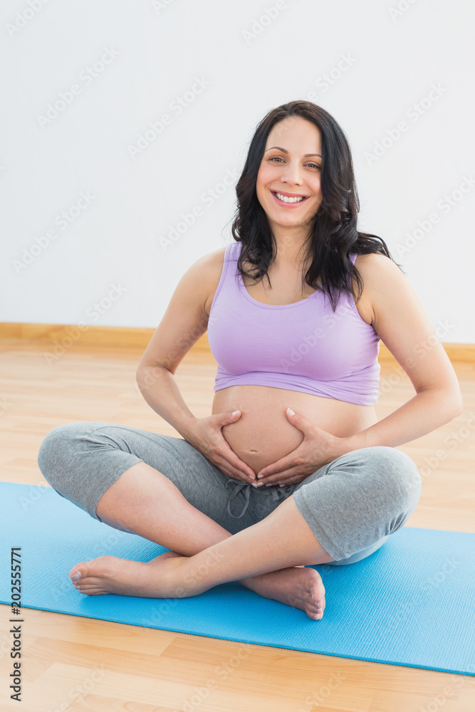 Pregnant brunette sitting on mat in touching her belly smiling at camera
