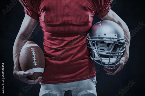 Midsection of American football player holding helmet and ball against grey background © vectorfusionart