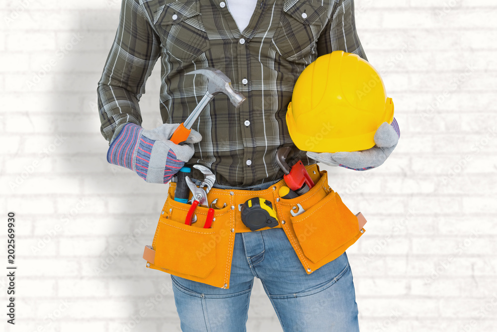Manual worker wearing tool belt while holding hammer and helmet against white wall Manual worker wearing tool belt while holding hammer and helmet against white wall