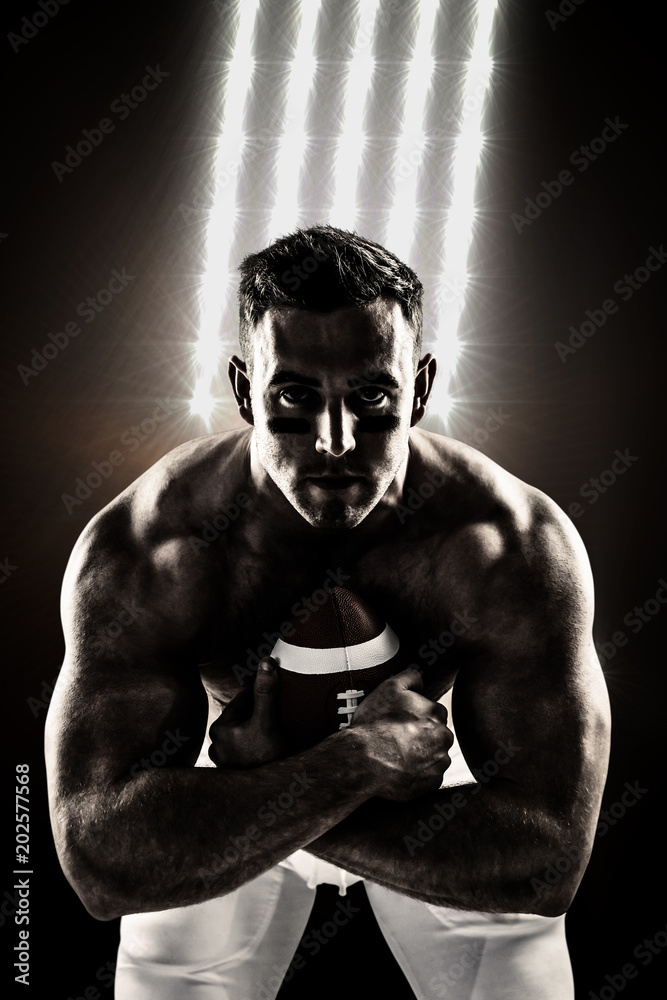 Shirtless American football player with ball against spotlights