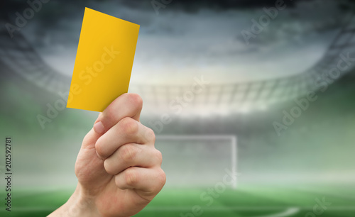 Hand holding up yellow card against football pitch in large stadium 