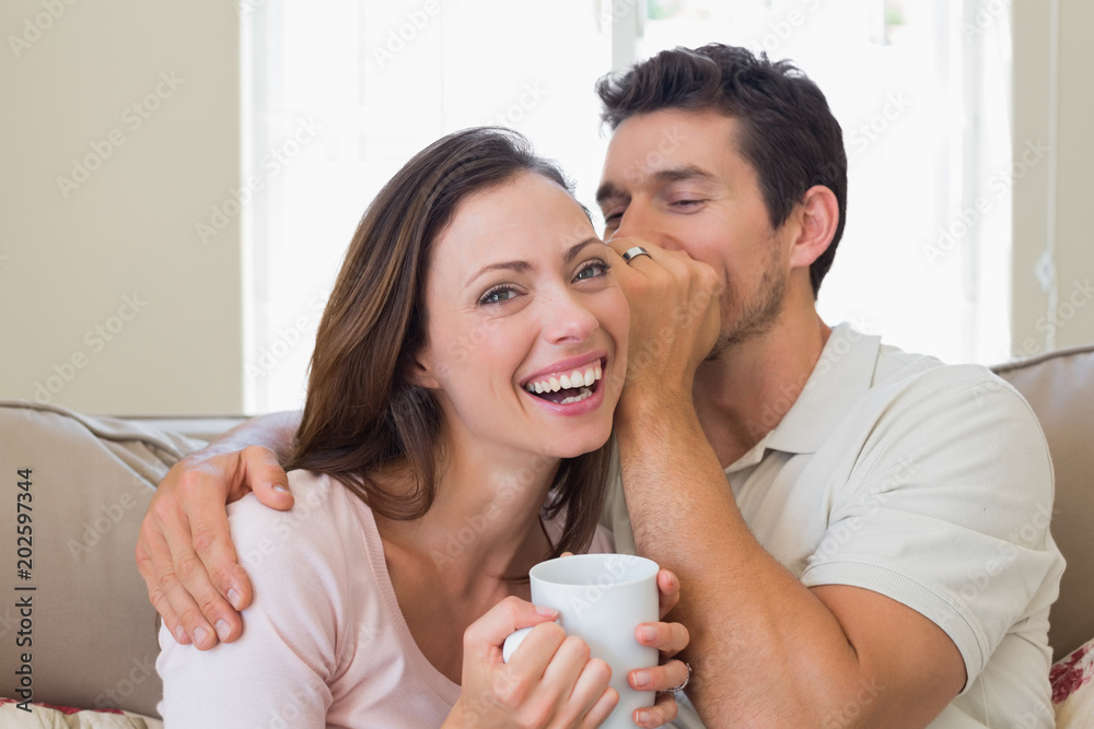 Man whispering secret into a cheerful womans ear in living room Man whispering secret into a cheerful womans ear in living room