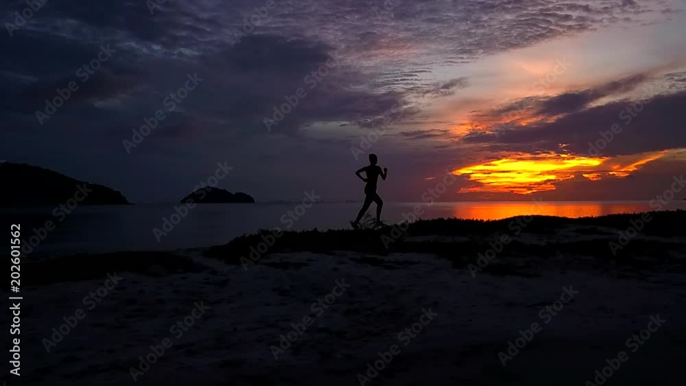 Tracking shot of woman silhouette jogging on the beach after sunset time.Fitness sport girl jogging fast training cardio near sea. Fly cam slow motion video