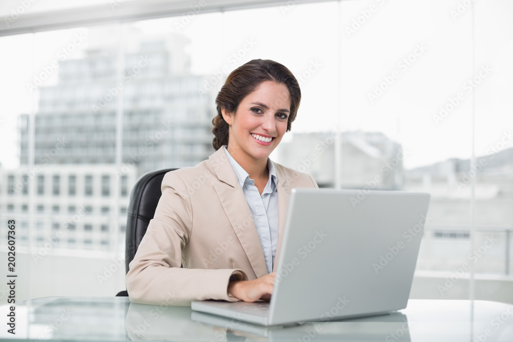 Happy businesswoman typing on laptop at her desk looking at camera Happy businesswoman typing on laptop at her desk looking at camera