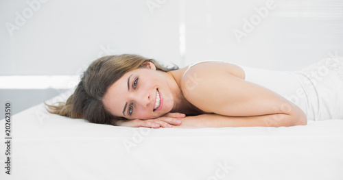Attractive brunette woman smiling cheerfully at camera lying on her bed