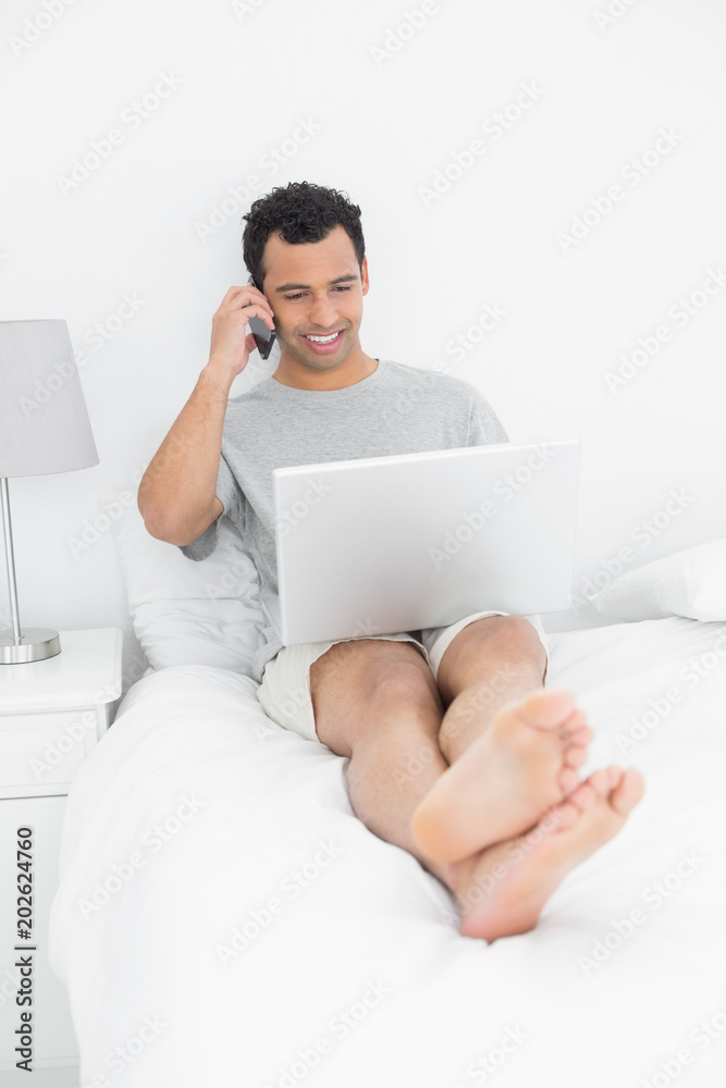 Casual smiling man using cellphone and laptop in bed Casual smiling man using cellphone and laptop in bed