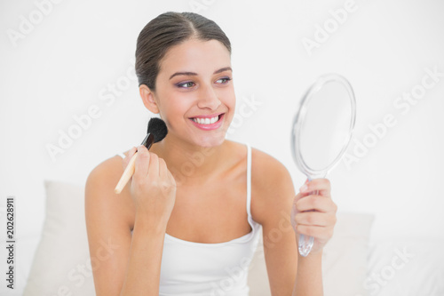 Smiling brown haired model in white pajamas applying powder on her face