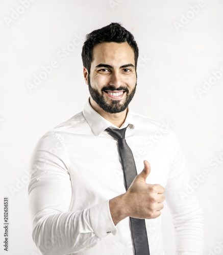 Portrait of bearded eastern man dressed in white shirt and black tie shows thumb up sign on isolated background