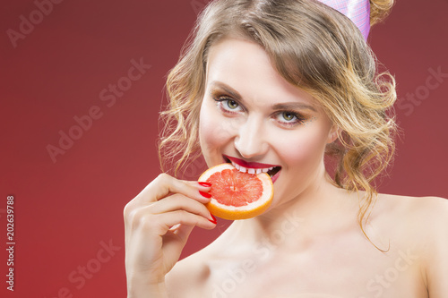 Fruit Ideas. Shy and Sexy Caucasian Blond Girl Biting Grapefruit Slice in Front. Against Red Background.