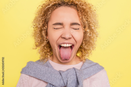 ortrait of a girl that has closed her eyes and showing her tounge outside of the mouth. She is very funny. Isolated on yellow bachground.