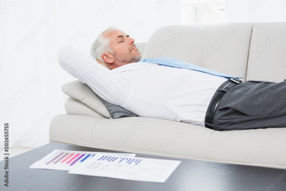 Fototapeta premium Businessman sleeping on sofa with graphs on table in living room
