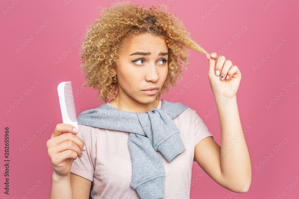 Young woman is looking at her hair. She is not satisfiet with its condition. Hair is blonde and curly. Girl is holding a white brush. Isolated on pink background. Young woman is looking at her hair. She is not satisfiet with its condition. Hair is blonde and curly. Girl is holding a white brush. Isolated on pink background.