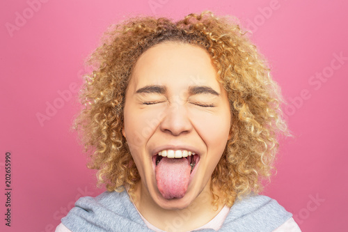 Close up of girl's face. She is showing her big tongue and keeping her eyes closed. This girl behaves like a child. Isolated on pink background.