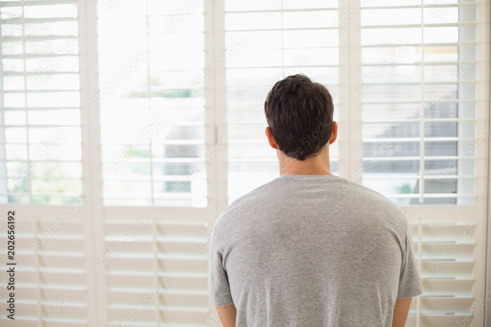 Fototapeta premium Rear view of man looking through window blinds at bright room