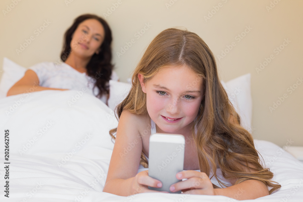 Girl using mobilephone on bed with mother in background Girl using mobilephone on bed with mother in background