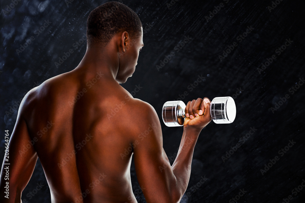 Rear view of a fit shirtless young man lifting dumbbell against black background