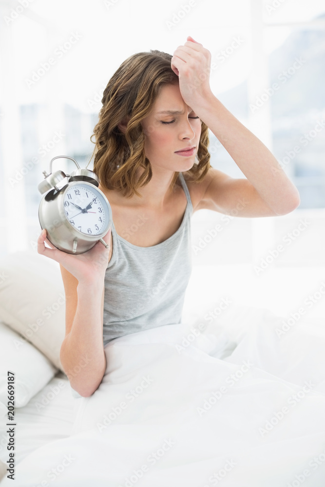 Annoyed young woman holding an alarm clock sitting on her bed