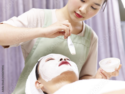 asian beautician applying facial mask on face of young woman