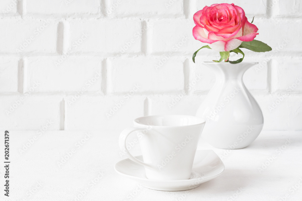 White cup and rose flower in a white vase on a light background of a brick wall in Skandmnavian minimalism a high key with a copy space White cup and rose flower in a white vase on a light background of a brick wall in Skandmnavian minimalism a high key with a copy space