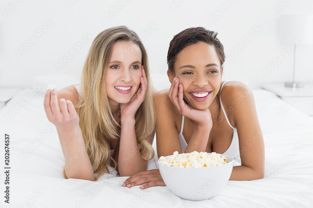 Cheerful female friends with popcorn bowl lying in bed Cheerful female friends with popcorn bowl lying in bed