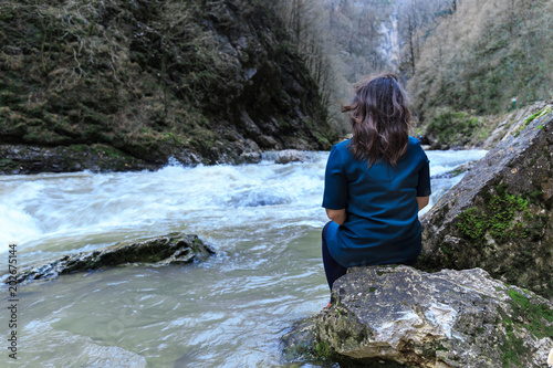 A girl sits with her back on the bank of a mountain river