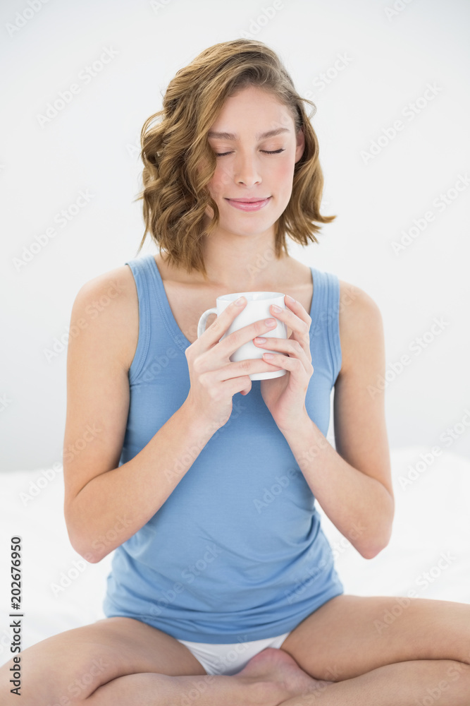 Content peaceful woman sitting on her bed holding white cup Content peaceful woman sitting on her bed holding white cup