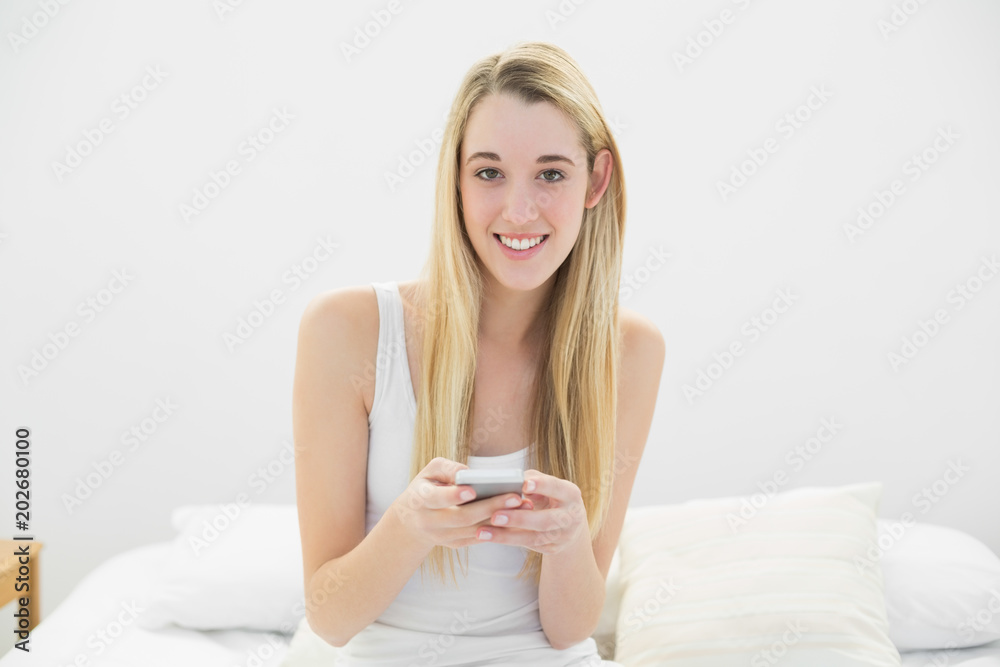 Lovely blonde woman texting with her smartphone sitting on her bed Lovely blonde woman texting with her smartphone sitting on her bed