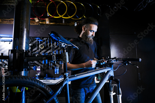 Young stylish bearded man with long hair Caucasian male mechanic worker at a bicycle workshop uses a tool for repairing a bicycle Bars with a hexagonal key photo