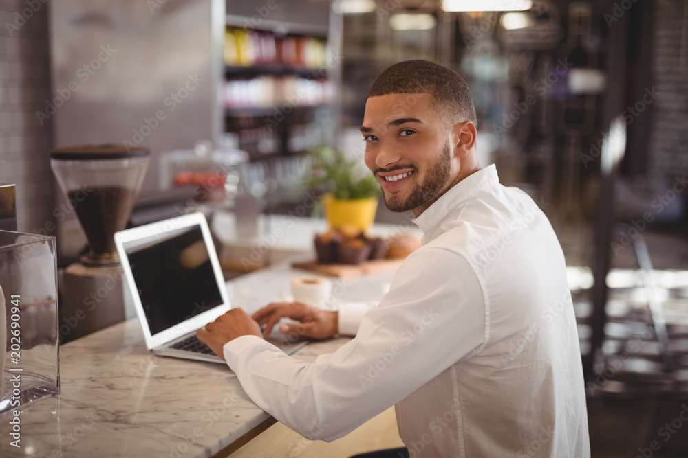 Portrait of smiling young male owner sitting with laptop at counter