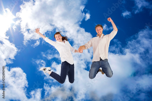 Couple jumping and holding hands against bright blue sky with clouds