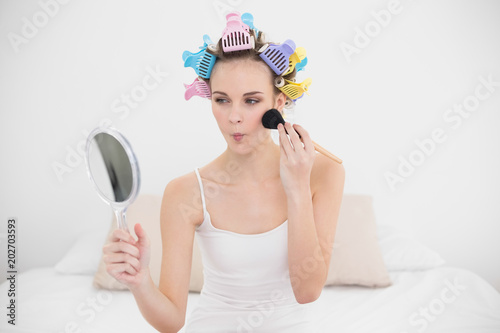 Pouting natural brown haired woman in hair curlers applying powder on her face