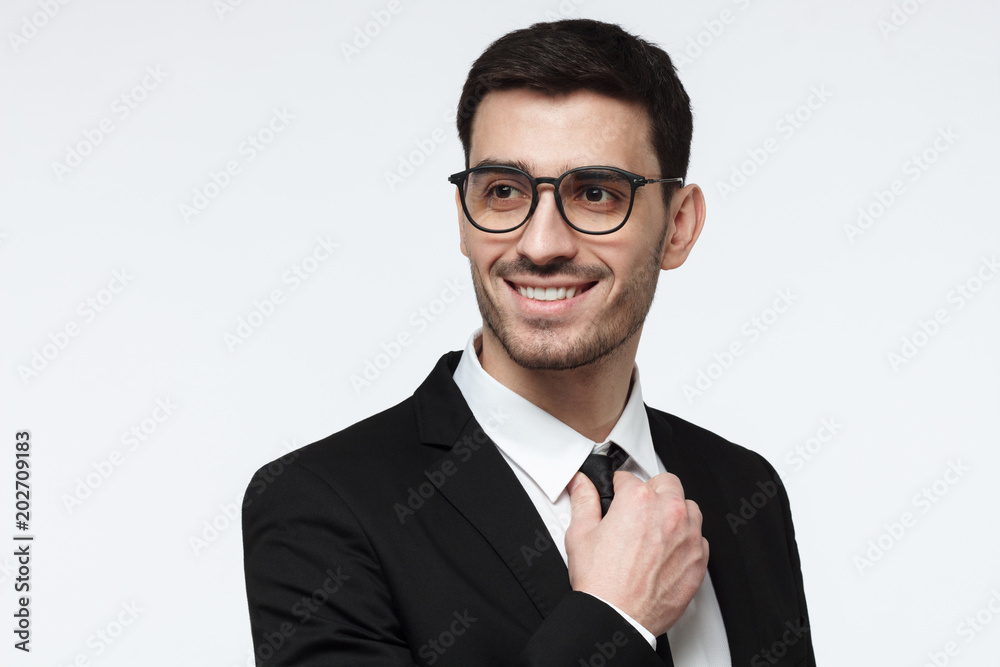 Closeup photo of handsome Caucasian businessman pictured isolated on grey background standing in black suit and eyeglasses looking aside with positive smile, feeling calm and ready to achieve goals Closeup photo of handsome Caucasian businessman pictured isolated on grey background standing in black suit and eyeglasses looking aside with positive smile, feeling calm and ready to achieve goals